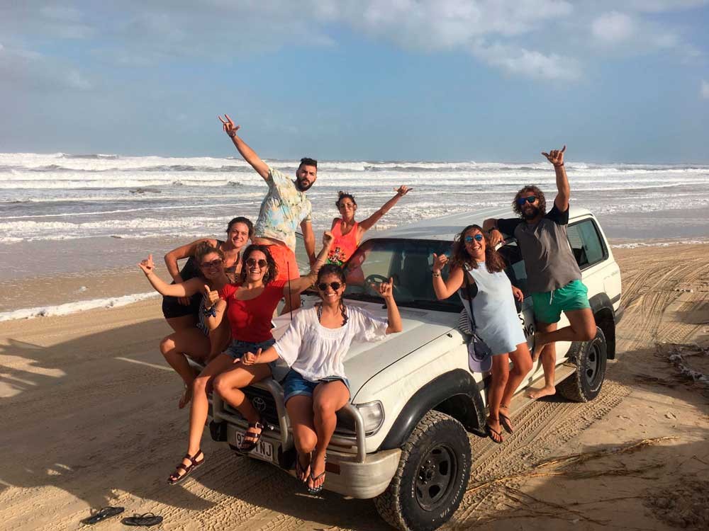 grupo de jóvenes encima de un jeep sonriendo a cámara y detrás la playa